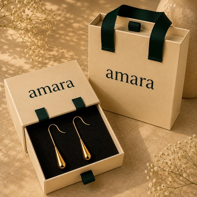 May include: A pair of gold-toned teardrop earrings with hook backings, presented in a beige sliding drawer box with a dark green ribbon pull. The box features the word amara printed in black. A matching beige gift bag with dark green handles is included.