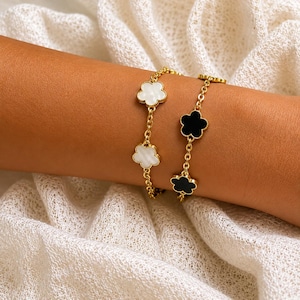 May include: Two gold-tone chain bracelets featuring flower-shaped charms. One bracelet displays white, iridescent floral motifs, while the other bracelet features solid black floral charms. Both pieces are constructed with delicate metal links.