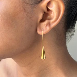 May include: A single gold-colored earring is shown, hanging from the ear. The earring has a long, thin wire that leads to a teardrop-shaped pendant. The pendant is smooth and shiny, reflecting light. The earring is a simple, elegant piece of jewelry.