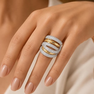 Puede incluir: Un anillo de múltiples bandas con filas alternas de metal pulido en tono dorado y piedras transparentes engastadas en pavé. El diseño consta de cinco bandas interconectadas que crean una apariencia ancha y superpuesta en el dedo, mostrando una mezcla de superficies metálicas lisas y texturas brillantes.