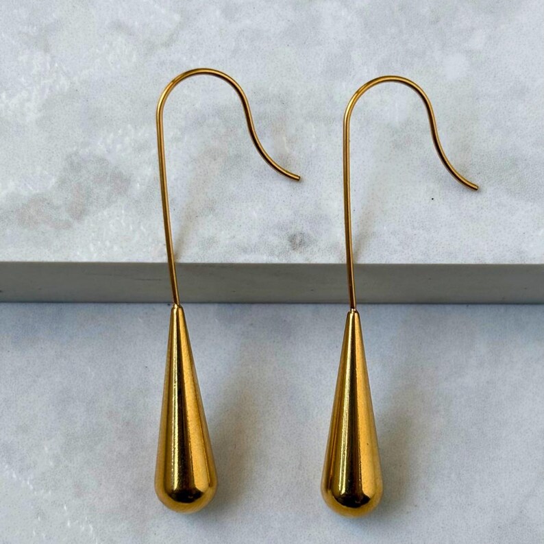 May include: A pair of gold-toned drop earrings featuring a smooth, elongated teardrop shape suspended from a curved wire hook. The metal surface has a polished, reflective finish that catches the light, creating a clean and simple aesthetic.