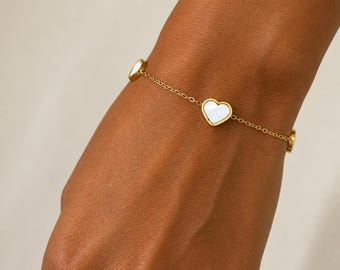 May include: A gold-toned chain bracelet featuring multiple heart-shaped charms with white, iridescent inlays. The delicate metal links connect each heart charm in a repeating pattern, creating a simple and refined piece of jewelry.