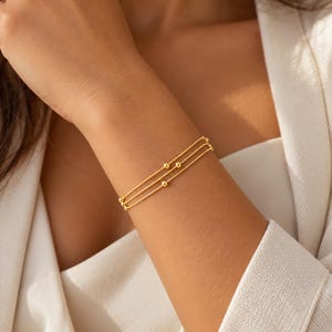 K&ouml;nnte beinhalten: Ein dreireihiges goldenes Armband mit d&uuml;nnen, zarten Ketten, die mit kleinen, gleichm&auml;&szlig;ig verteilten goldenen Perlen verziert sind. Das Armband besteht aus drei parallelen Str&auml;ngen, die das Handgelenk umschlie&szlig;en und einen geschichteten Look mit polierter Oberfl&auml;che erzeugen.
