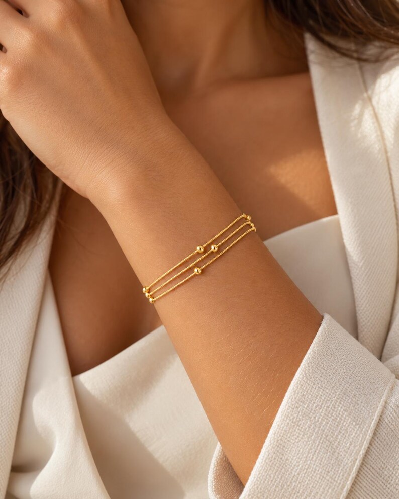 K&ouml;nnte beinhalten: Ein dreireihiges goldenes Armband mit d&uuml;nnen, zarten Ketten, die mit kleinen, gleichm&auml;&szlig;ig verteilten goldenen Perlen verziert sind. Das Armband besteht aus drei parallelen Str&auml;ngen, die das Handgelenk umschlie&szlig;en und einen geschichteten Look mit polierter Oberfl&auml;che erzeugen.