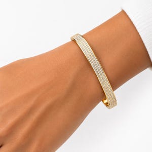 May include: A gold-toned bangle bracelet featuring three parallel rows of small, clear, sparkling stones embedded along the outer surface. The bracelet has a smooth, polished finish and a hinged design, providing a secure fit around the wrist.