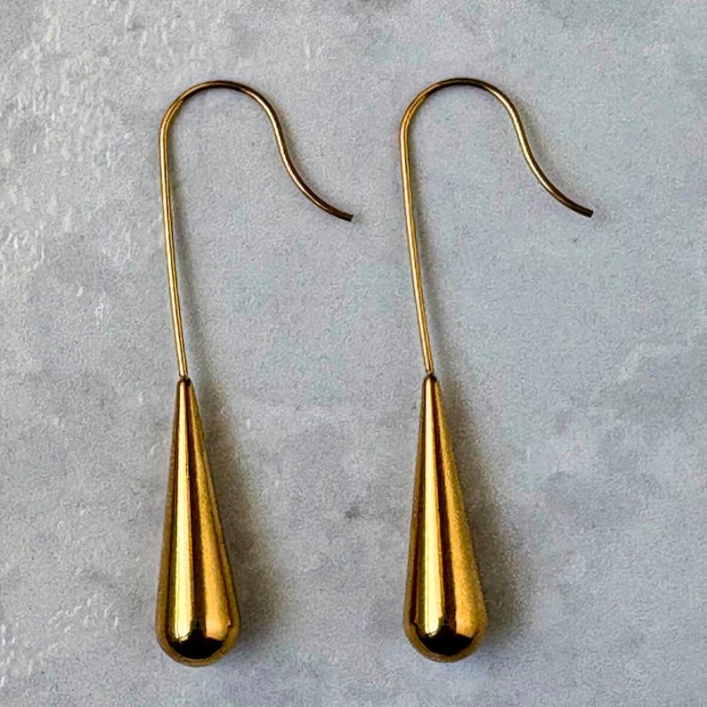 May include: A pair of gold-toned drop earrings featuring a smooth, polished teardrop-shaped pendant suspended from a curved wire hook. The metal surface is reflective and solid, showcasing a simple, elongated design that hangs vertically.