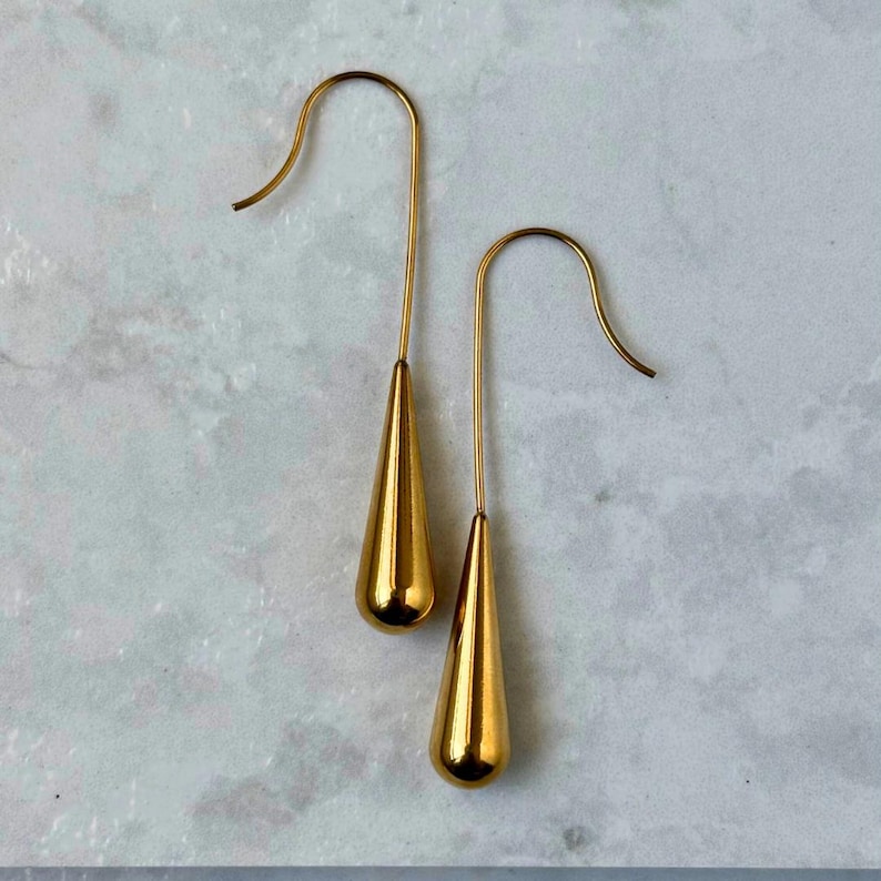 May include: A pair of gold-toned drop earrings featuring smooth, elongated teardrop-shaped pendants suspended from curved wire hooks. The metal surface has a polished, reflective finish that catches the light, creating a simple and clean aesthetic.