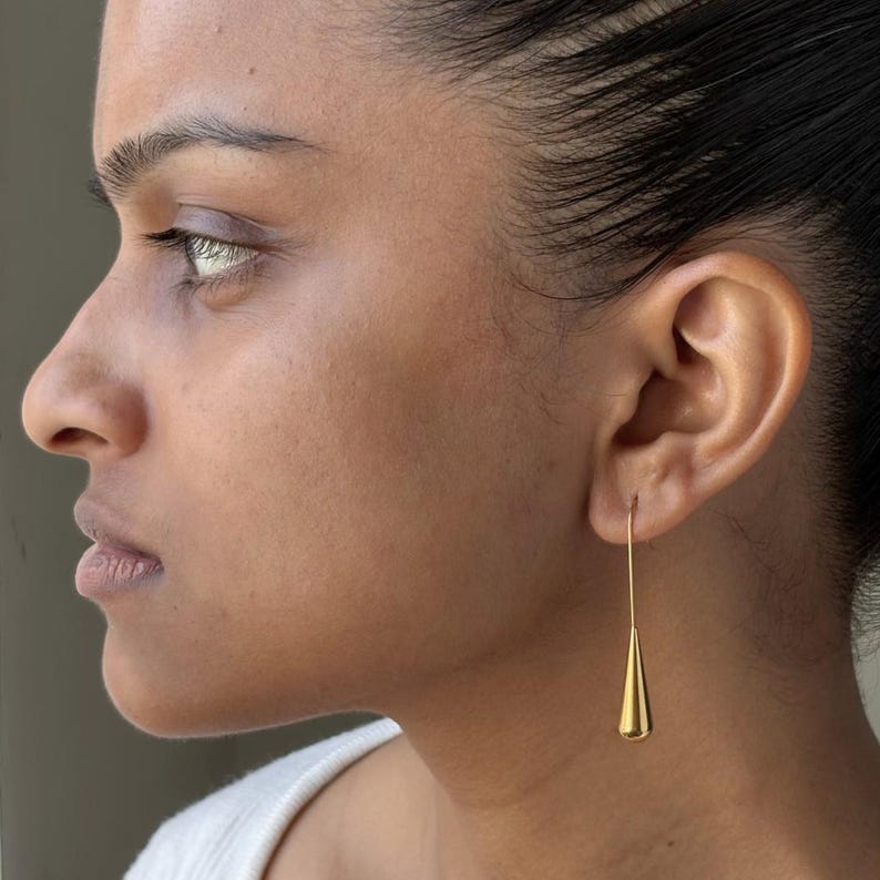 May include: A gold-toned metal earring featuring a long, thin wire post that connects to a smooth, polished, teardrop-shaped pendant. The earring hangs vertically from the earlobe, showcasing a minimalist design with a reflective, metallic finish.