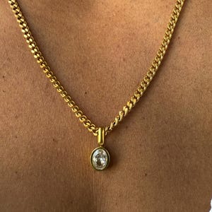 May include: A gold-coloured chain necklace with a pendant. The pendant has a clear, oval-shaped stone set in a gold bezel. The chain is a thick, curb style, and the necklace is shown against a skin-toned background.