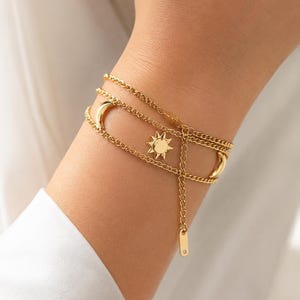 May include: Gold-coloured layered bracelet with sun and crescent moon charms. The bracelet has multiple delicate chains and a small tag. The jewellery is worn on a wrist, with a white shirt sleeve in the background.