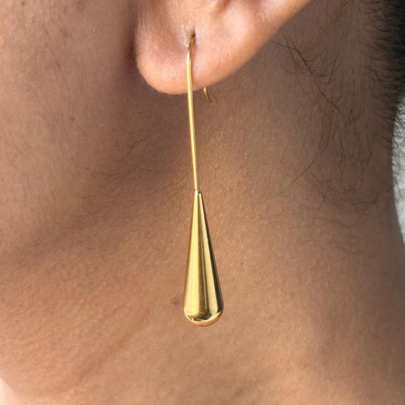 May include: A gold-toned drop earring featuring a long, slender wire hook that connects to a smooth, polished, teardrop-shaped pendant. The metal surface has a reflective, metallic finish, creating a clean and simple aesthetic for this jewellery piece.