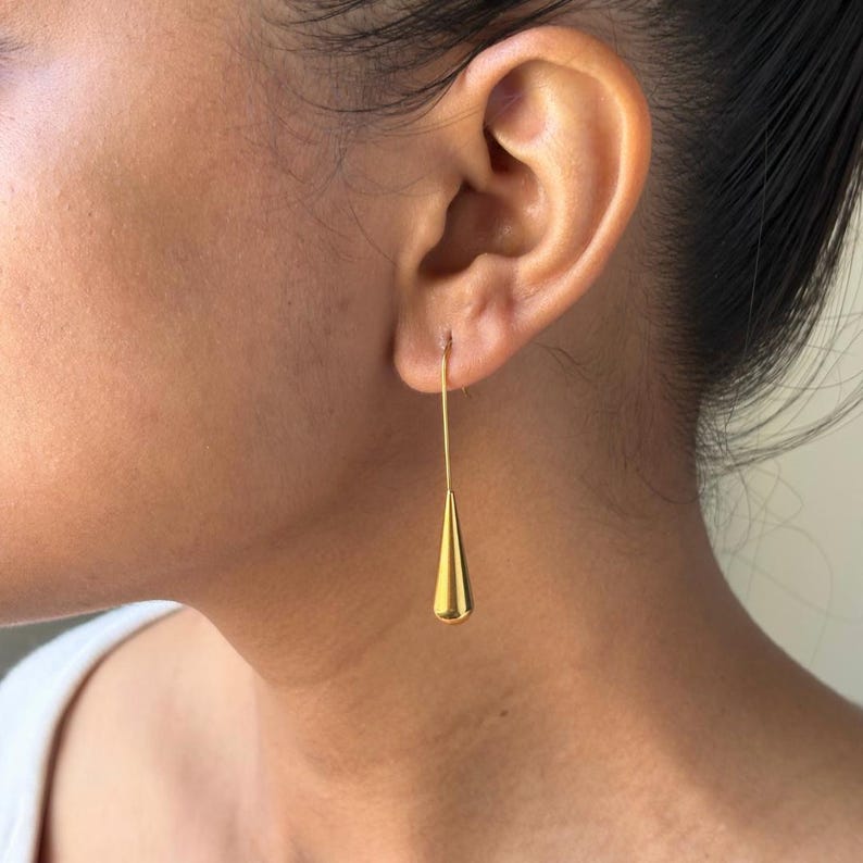 May include: A gold-toned drop earring featuring a long, thin wire stem attached to a smooth, polished teardrop-shaped pendant. The earring hangs vertically from the earlobe, showcasing a reflective metallic finish and a simple, elongated design.