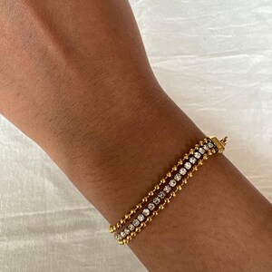 Könnte beinhalten: Goldfarbenes Armband mit drei Strängen. Die oberen und unteren Stränge haben kleine, runde Perlen, während der mittlere Strang mit einer Reihe klarer Kristalle besetzt ist. Das Armband wird mit einem kleinen Verschluss geschlossen.
