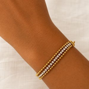 May include: A gold-toned multi-strand bracelet featuring two outer chains of small polished beads and a central row of clear, round-cut sparkling stones. The bracelet is secured with a rectangular gold-toned clasp at the end of the three strands.