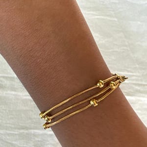 K&ouml;nnte beinhalten: Ein goldfarbenes Armband bestehend aus drei d&uuml;nnen, flexiblen Schlangenketten, die &uuml;bereinander liegen. Das Design umfasst kleine, runde, polierte Goldperlen, die gleichm&auml;&szlig;ig entlang der Ketten verteilt sind und mit einem Metallverschluss enden.