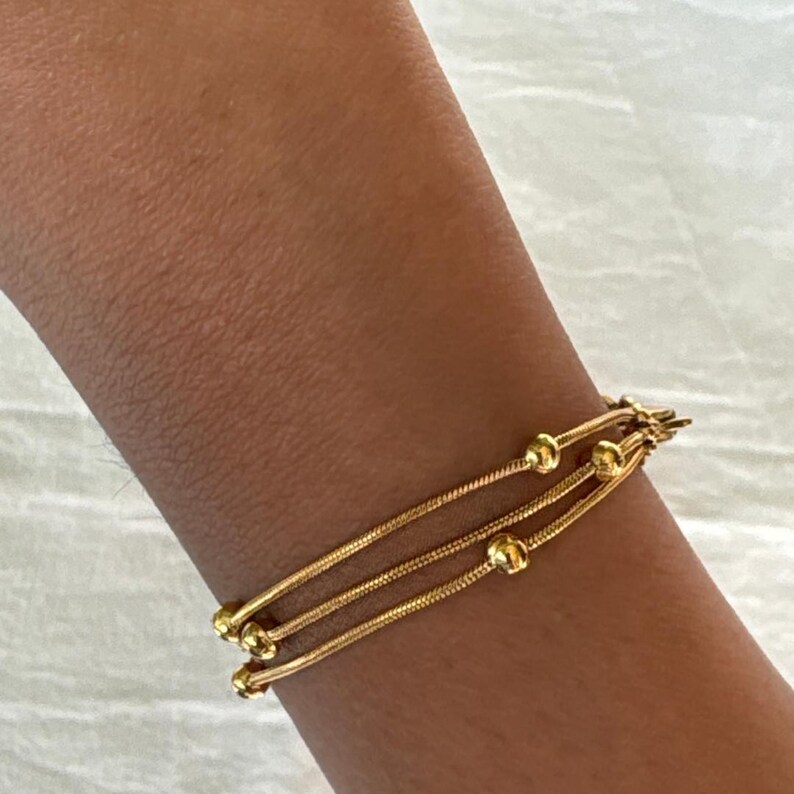 K&ouml;nnte beinhalten: Ein goldfarbenes Armband bestehend aus drei d&uuml;nnen, flexiblen Schlangenketten, die &uuml;bereinander liegen. Das Design umfasst kleine, runde, polierte Goldperlen, die gleichm&auml;&szlig;ig entlang der Ketten verteilt sind und mit einem Metallverschluss enden.