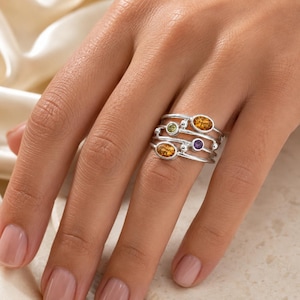 May include: A multi-band silver ring featuring four distinct gemstone settings. The ring includes two oval-shaped amber-colored stones, one small round green stone, and one small round purple stone, all set within a polished silver metal frame.