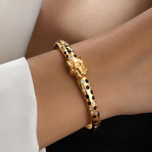 May include: Gold-coloured bangle bracelet with a leopard head clasp. The bracelet has a black and white polka dot pattern. The leopard head is detailed with textured fur and facial features. A stylish piece of jewellery.