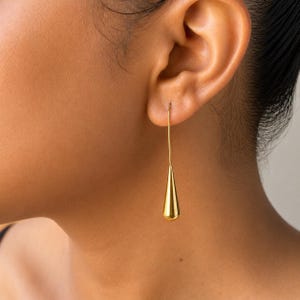 May include: A gold-toned drop earring featuring a long, thin wire stem that connects to a smooth, polished, teardrop-shaped pendant. The earring hangs vertically from the earlobe, showcasing a reflective metallic finish and a sleek, elongated design.