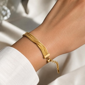 Puede incluir: Una pulsera dorada con múltiples hebras se muestra en una muñeca. La pulsera tiene un cierre rectangular y una cadena ajustable. La muñeca está contra un fondo blanco, y la persona lleva una camisa blanca.