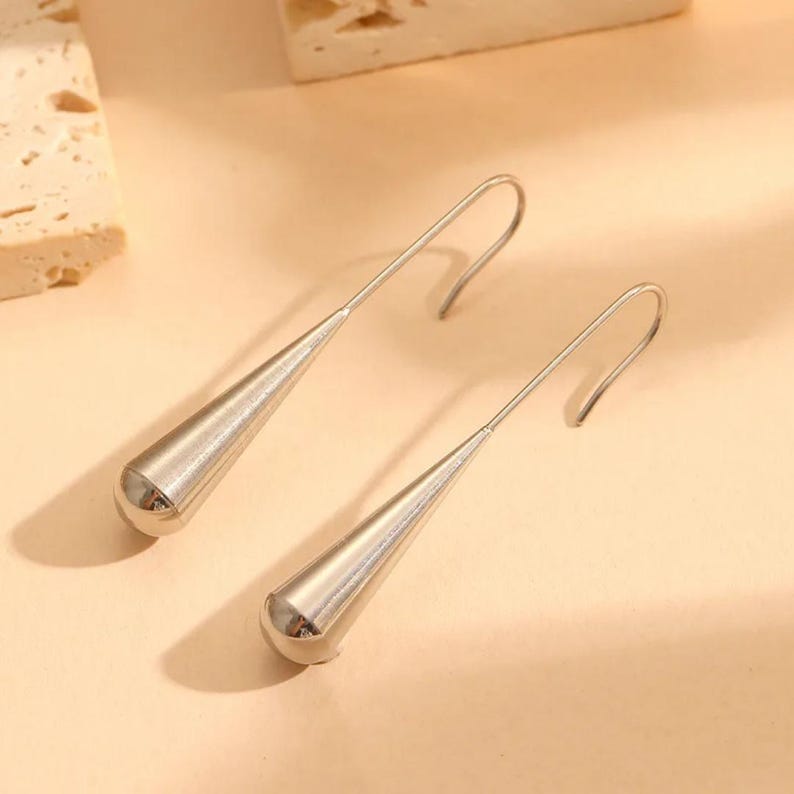 May include: A pair of silver-toned drop earrings featuring a smooth, elongated teardrop or cone-shaped pendant suspended from a curved hook wire. The metal surface has a polished, reflective finish that catches the light, creating a sleek appearance.