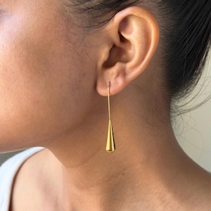 May include: A gold-toned drop earring featuring a long, thin wire stem that connects to a smooth, polished teardrop-shaped pendant. The earring hangs vertically from the earlobe, showcasing a reflective metallic finish and a clean, simple design.