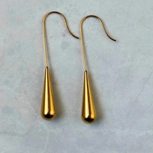 May include: A pair of gold-toned drop earrings featuring a smooth, polished teardrop-shaped pendant suspended from a curved wire hook. The earrings have a reflective metallic finish and a simple, elongated design that hangs vertically.