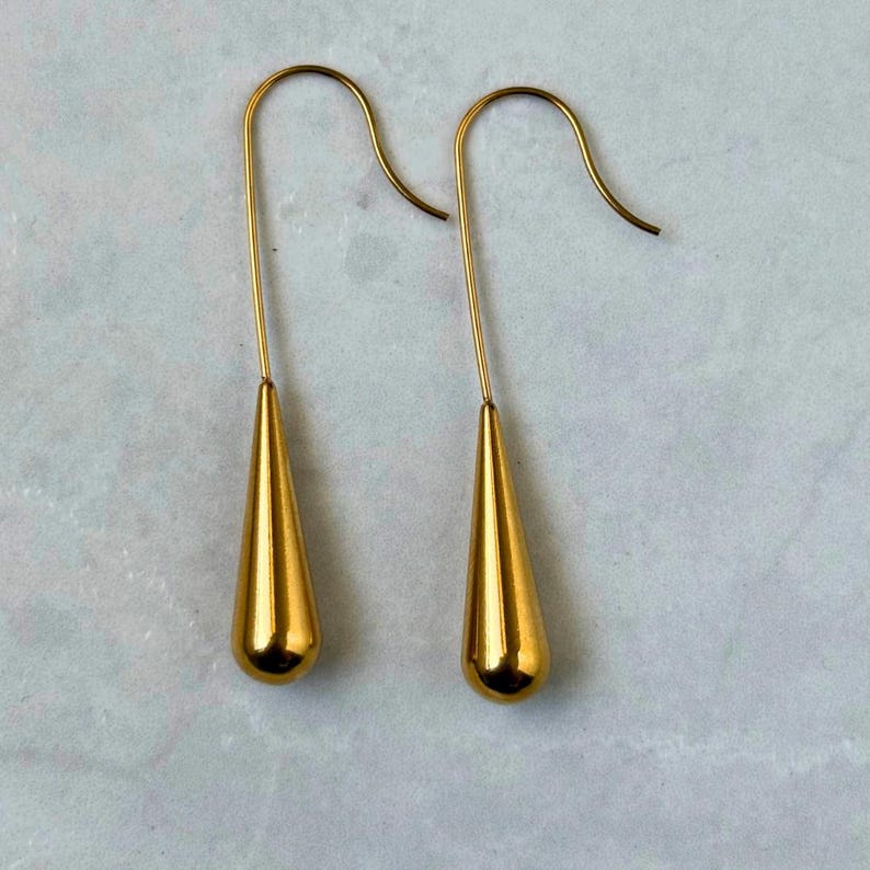 May include: A pair of gold-toned drop earrings featuring a smooth, polished teardrop-shaped pendant suspended from a curved wire hook. The earrings have a reflective metallic finish and a simple, elongated design that hangs vertically.