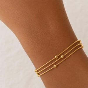 K&ouml;nnte beinhalten: Ein goldfarbenes Armband bestehend aus drei d&uuml;nnen, flexiblen Schlangenketten, die &uuml;bereinander liegen. Jede Kette ist mit kleinen, glatten, runden Goldperlen verziert, die entlang der Metallstr&auml;nge verteilt sind und ein zartes Aussehen bieten.