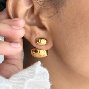May include: A close-up of gold-colored, rounded stud earrings. The earrings are smooth and reflective, with a polished finish. The earrings are on a person's ear.