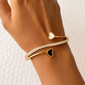 May include: A gold-tone open cuff bracelet featuring a wavy band encrusted with small clear crystals. The two ends of the bracelet terminate in heart-shaped charms, one inlaid with a white stone and the other with a black stone.