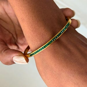 May include: A gold-coloured bangle bracelet featuring a line of emerald-green gemstones. The bracelet is held by a hand with a light skin tone. The gemstones are rectangular and closely set.