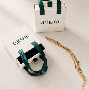 K&ouml;nnte beinhalten: Zwei quadratische wei&szlig;e Geschenkboxen mit dunkelgr&uuml;nen Bandgriffen und dem in Gr&uuml;n gedruckten Text amara. Eine Box ist geschlossen, die andere ist ge&ouml;ffnet und zeigt ein schwarzes Inneres. Ein goldfarbenes mehrstr&auml;ngiges Kettenarmband mit kleinen Perlen.
