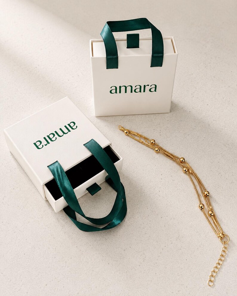 K&ouml;nnte beinhalten: Zwei quadratische wei&szlig;e Geschenkboxen mit dunkelgr&uuml;nen Bandgriffen und dem in Gr&uuml;n gedruckten Text amara. Eine Box ist geschlossen, die andere ist ge&ouml;ffnet und zeigt ein schwarzes Inneres. Ein goldfarbenes mehrstr&auml;ngiges Kettenarmband mit kleinen Perlen.