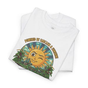 May include: Two white t-shirts are stacked, with the top shirt featuring a colorful graphic. The graphic includes a winking sun, windmills, and the text "Powered by Sarcasm & Sunshine" in a circular design. The design has a retro, cartoonish style.