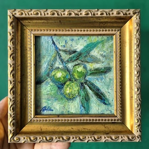 May include: A small, framed oil painting of green olives on a branch. The artwork features a gold-colored ornate frame and a textured, impressionistic style. The painting's background is a mix of blue and green hues.