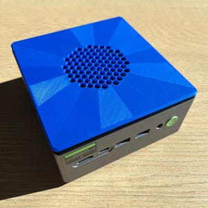 May include: A small, silver computer with a bright blue top. The blue top has a geometric design and a circular vent. The computer has several ports and a power button. A sticker with text is on the side.