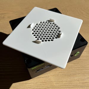 May include: A white square cover with a honeycomb-patterned vent is placed on top of a black rectangular device. The cover has four small tabs. The device has a green sticker and a slot for a card.