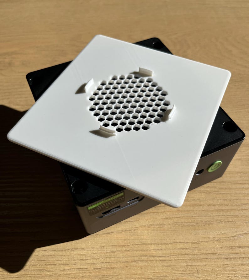 May include: A white square cover with a honeycomb-patterned vent is placed on top of a black rectangular device. The cover has four small tabs. The device has a green sticker and a slot for a card.