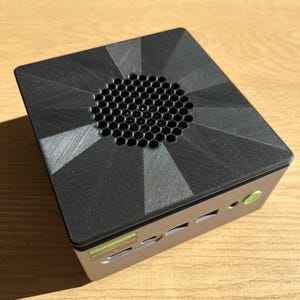 May include: A small, black computer case with a geometric design on the top. The case has a circular vent with a honeycomb pattern and several ports on the side. A green power button is visible.