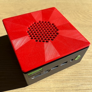 May include: A small, square computer with a red, geometric top featuring a honeycomb-patterned vent. The sides are dark gray, with several ports and a green power button. The device sits on a light-colored wooden surface.