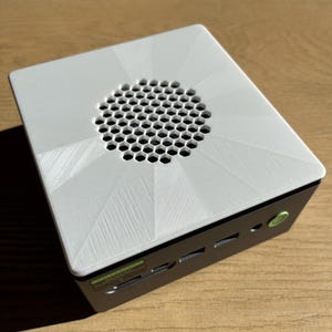 May include: A small, white computer case with a geometric design on the top. The top has a circular honeycomb vent. The case is black on the sides with a green power button and various ports.