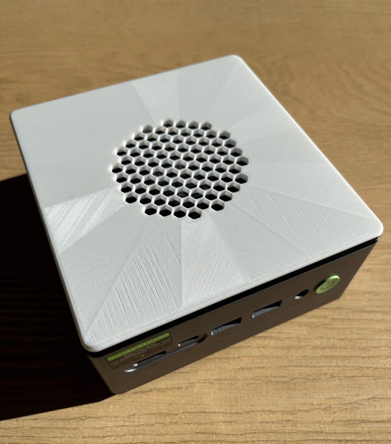May include: A small, white computer case with a geometric design on the top. The top has a circular honeycomb vent. The case is black on the sides with a green power button and various ports.