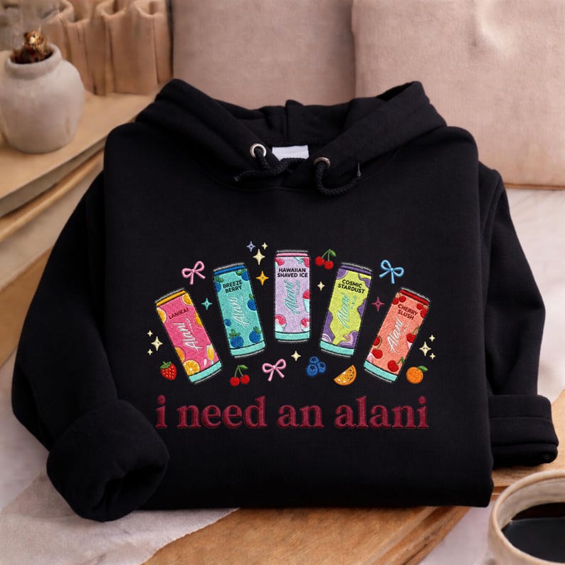 A black hoodie with a colorful graphic design featuring five cans of flavored waters and the text "I need an alani" printed on it.