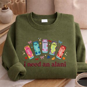A green sweatshirt with a colorful graphic design featuring five different flavored drinks and the text "I need an alani" printed on it.