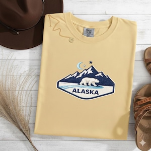 May include: A light yellow t-shirt with an embroidered Alaska design. The design features a polar bear, mountains, a crescent moon, and the word "Alaska". The shirt has a crew neck and short sleeves.