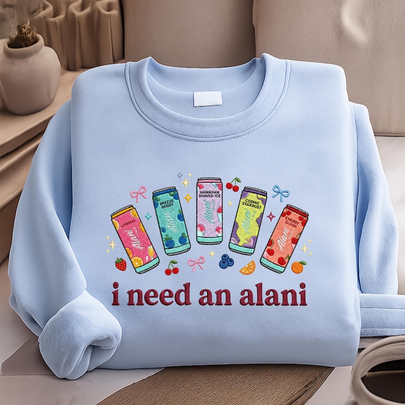 A light blue sweatshirt with a graphic design featuring five colorful cans of soda and the text "I need an alani" printed on it.
