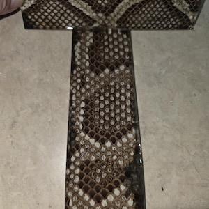 May include: A rectangular piece of material with a snakeskin pattern in shades of brown, tan, and white. The material is held by a hand, and the pattern is visible on the surface.