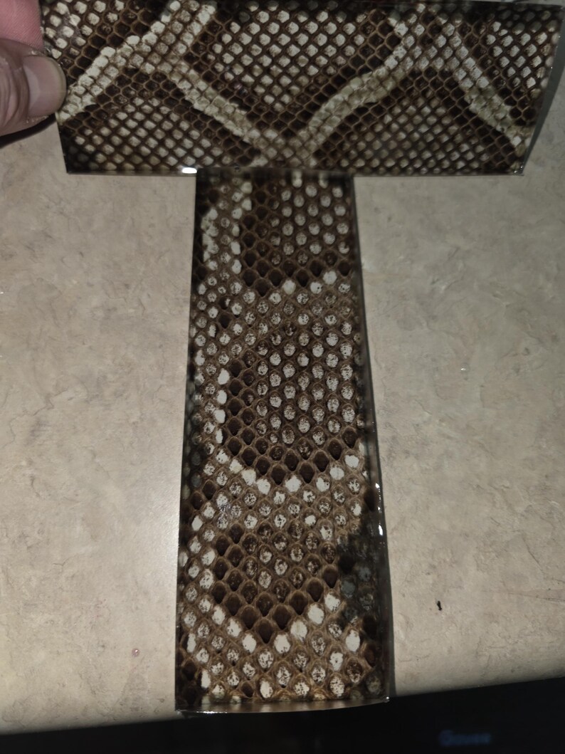 Pode incluir: Uma pe&ccedil;a retangular de material com um padr&atilde;o de pele de cobra em tons de castanho, bege e branco. O material &eacute; segurado por uma m&atilde;o, e o padr&atilde;o &eacute; vis&iacute;vel na superf&iacute;cie.