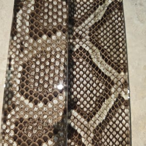 Pode incluir: Duas pe&ccedil;as retangulares com um padr&atilde;o de pele de cobra. O padr&atilde;o apresenta uma mistura de escamas marrons, bege e brancas, criando uma textura realista de pele de r&eacute;ptil. As pe&ccedil;as t&ecirc;m um acabamento brilhante.